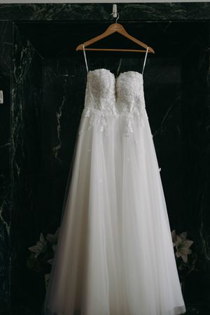Elegant white wedding dress with lace bodice and tulle skirt hanging on wooden hanger against dark wall.