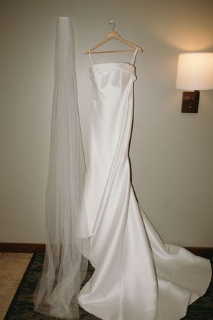 A white wedding dress on a wooden hanger displayed against a neutral wall with soft lighting and flowing veil.
