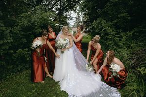 A bride in a white lace gown stands with bridesmaids wearing rust-colored dresses among green foliage outdoors.