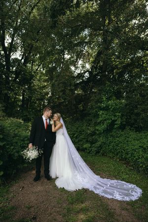 A wedding couple stands together on a woodland path with a flowing white veil trailing behind the bride.