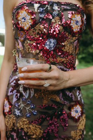 Close up of hands holding drink wearing a floral sequined strapless dress with pink, gold and purple embellishments.