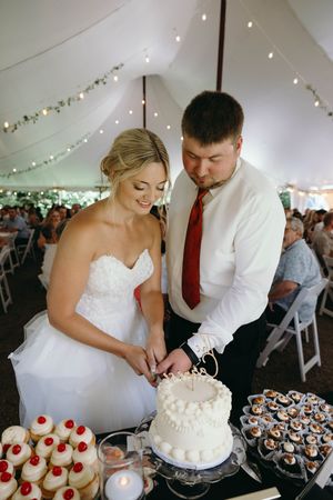 A couple cuts their white wedding cake together at a dessert table with cupcakes and string lights overhead in a tent.