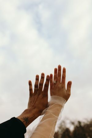 Two hands with rings reach up towards a cloudy sky, one wearing black and one wearing white sleeves.