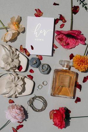 Flatlay photography with roses, perfume bottle, rings, vows card and decorative elements on white background.
