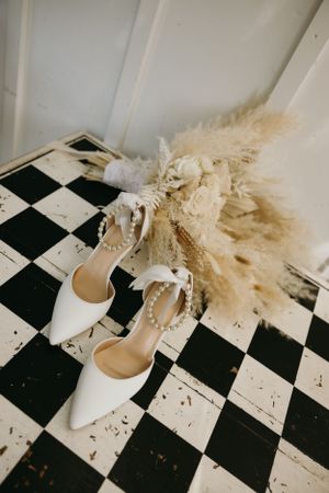 White bridal shoes and dried floral arrangement on checkered floor.