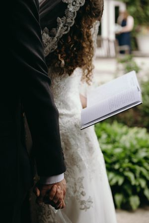 Close-up of a couple during wedding ceremony with bride in lace dress holding open book against garden background.