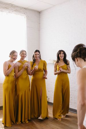 Bridesmaids in matching golden yellow dresses smile and laugh together in a bright white room.