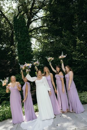 A group of bridesmaids in lilac purple dresses and a bride in white celebrate joyfully together in an outdoor garden setting.