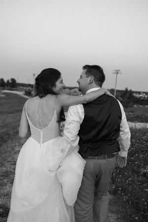 A couple in wedding attire embracing while walking together in a rustic outdoor setting at dusk.