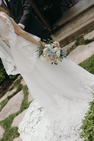 A delicate white lace wedding dress with flowing train and pastel blue and pink floral bouquet shown against stone path.