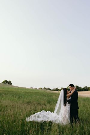 A romantic wedding photo of a couple embracing in a serene grassy meadow at dusk, with a flowing white veil and dress.