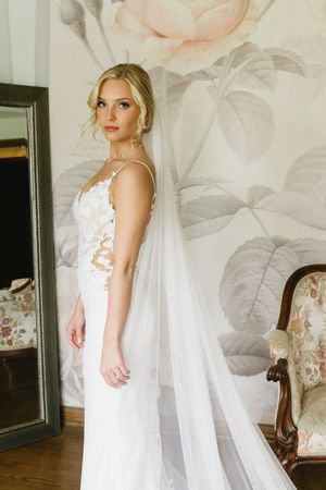 A bride in a white lace wedding gown and long veil poses against a floral wallpaper background with antique furniture.