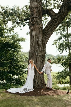 A couple in wedding attire stands on opposite sides of a large tree trunk during their first look photo moment outdoors.