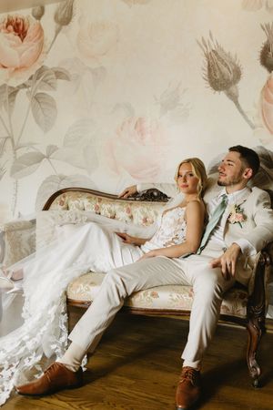 Elegant couple relaxing on vintage settee against floral wallpaper in romantic wedding photoshoot.