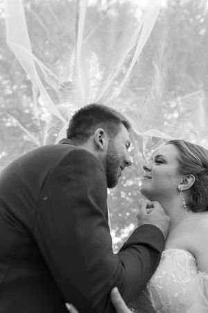 An artistic black and white close-up of newlyweds with flowing veil in natural lighting.