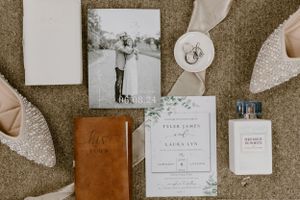 Wedding stationery flatlay with invitation, leather journal, perfume and shoes on carpet.