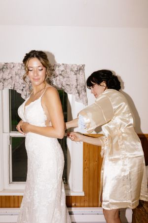 Behind the scenes of wedding preparations with final dress adjustments in vintage-style lighting.
