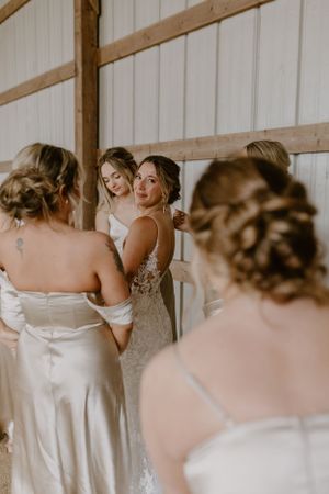 Group gathered for wedding preparations in a rustic barn setting with soft lighting and elegant white dresses.