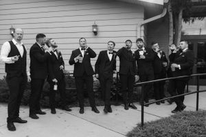 Groomsmen lined up outdoors wearing formal black attire and holding drinks for a wedding celebration.