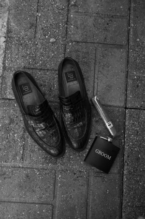 Black dress shoes and groom's flask arranged on concrete surface in monochrome style.