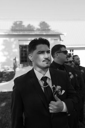 A black and white photo showing a groom and groomsmen wearing formal suits with boutonnières at an outdoor wedding.