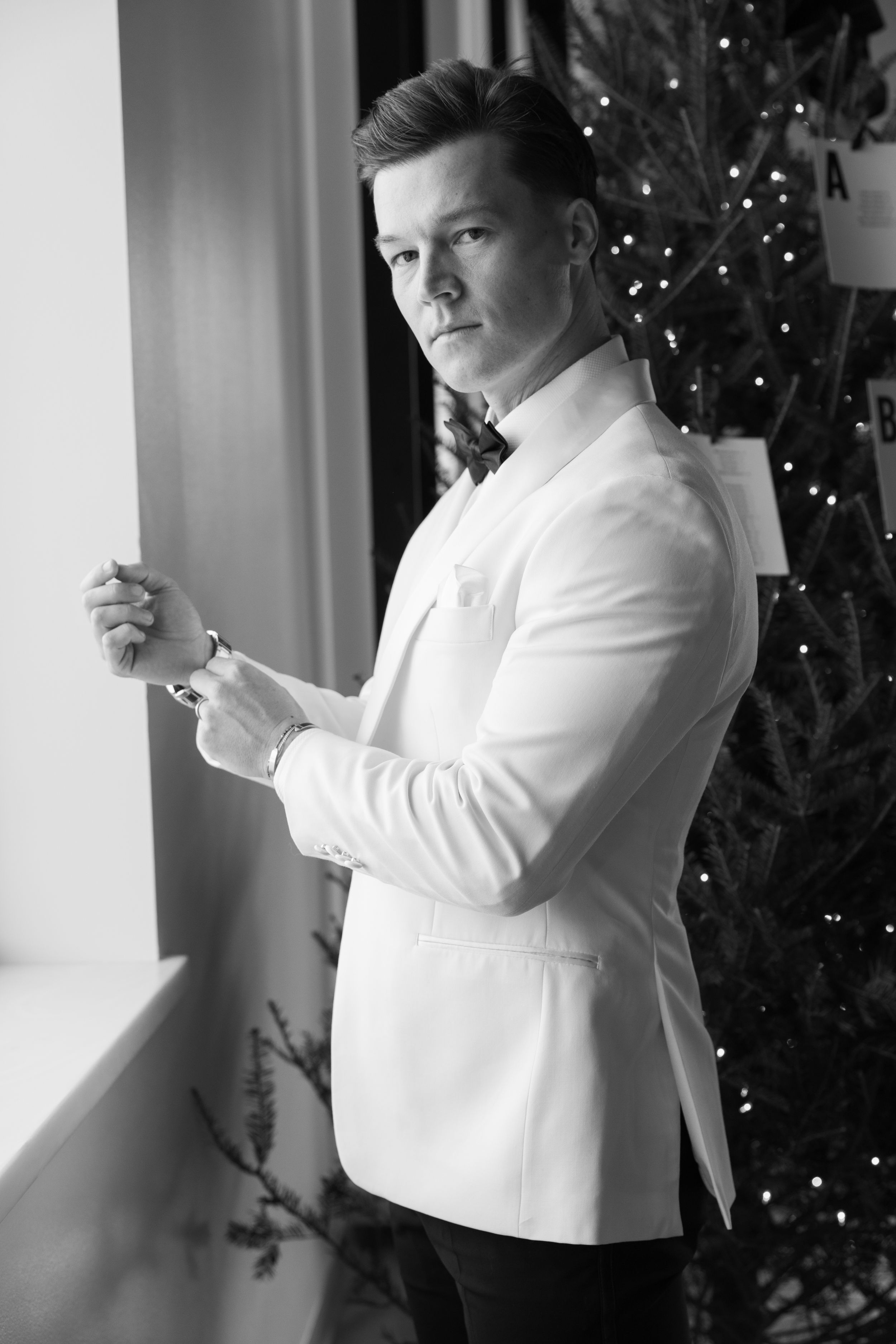 Black and white photo of person in white dinner jacket adjusting cufflinks by window with Christmas tree in background.