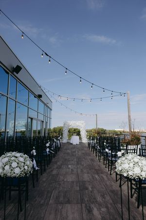 Outdoor rooftop venue with string lights and white floral arrangements along a modern glass-walled space at sunset.