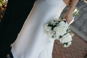 A bride in a white wedding dress holds a classic white bouquet while standing on a brick pathway.