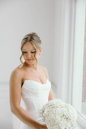 Bride in white strapless wedding dress holds elegant white hydrangea bouquet while looking down in bright window light.