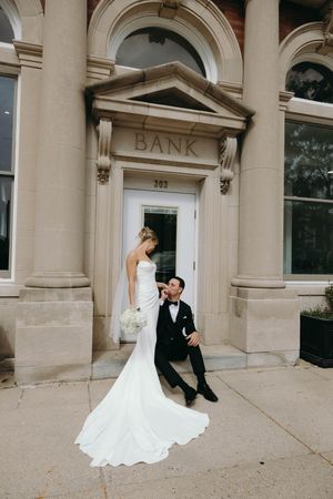 A romantic wedding photo series showing a couple embracing in front of a historic bank building's stone facade.