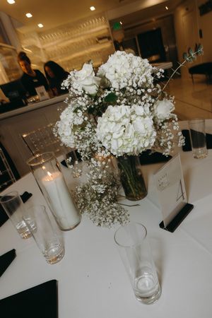 White floral centerpieces in glass vases decorate a minimalist table setting with soft overhead lighting.