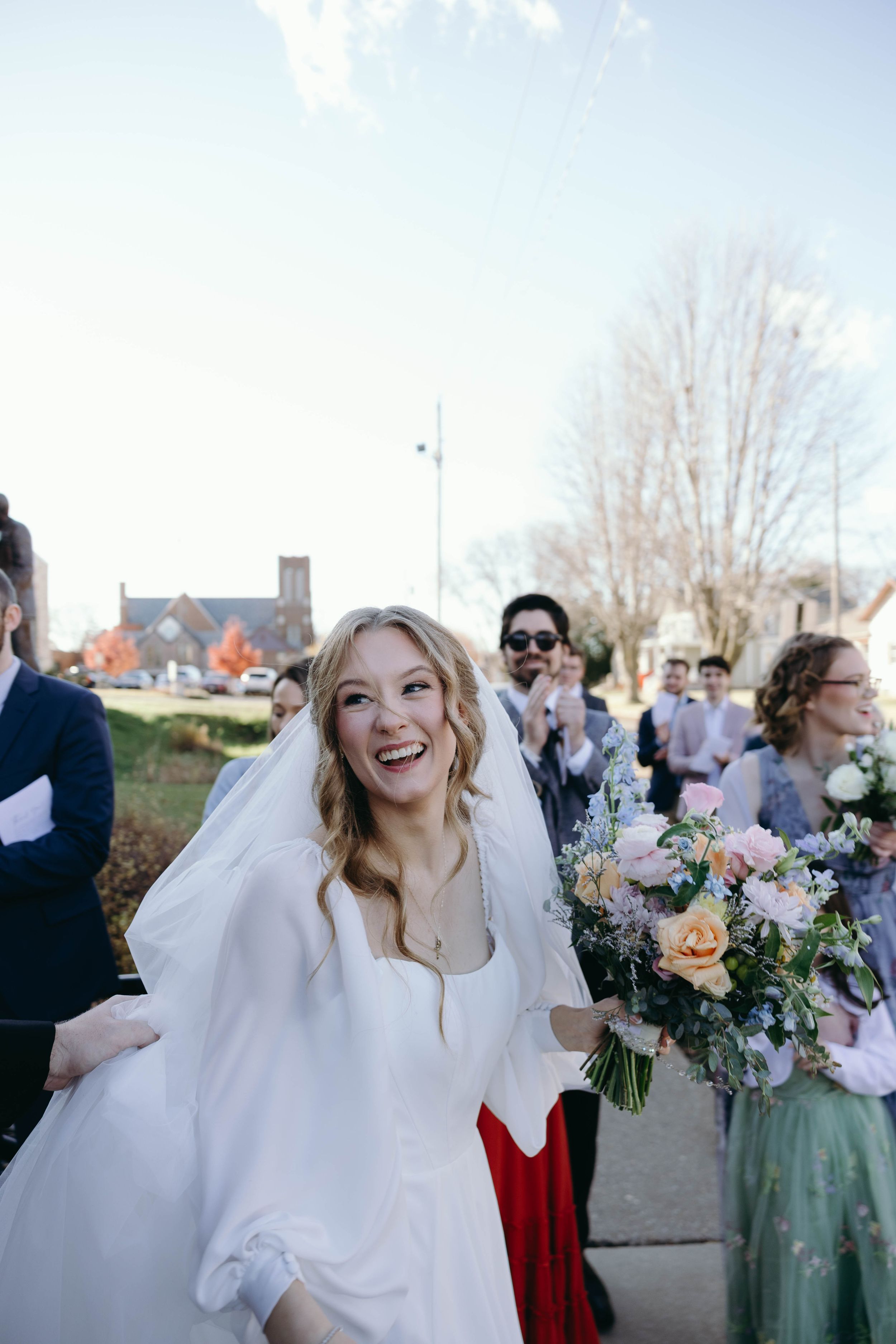 A bride in a white dress laughs joyfully while holding a floral bouquet outdoors at a wedding celebration.
