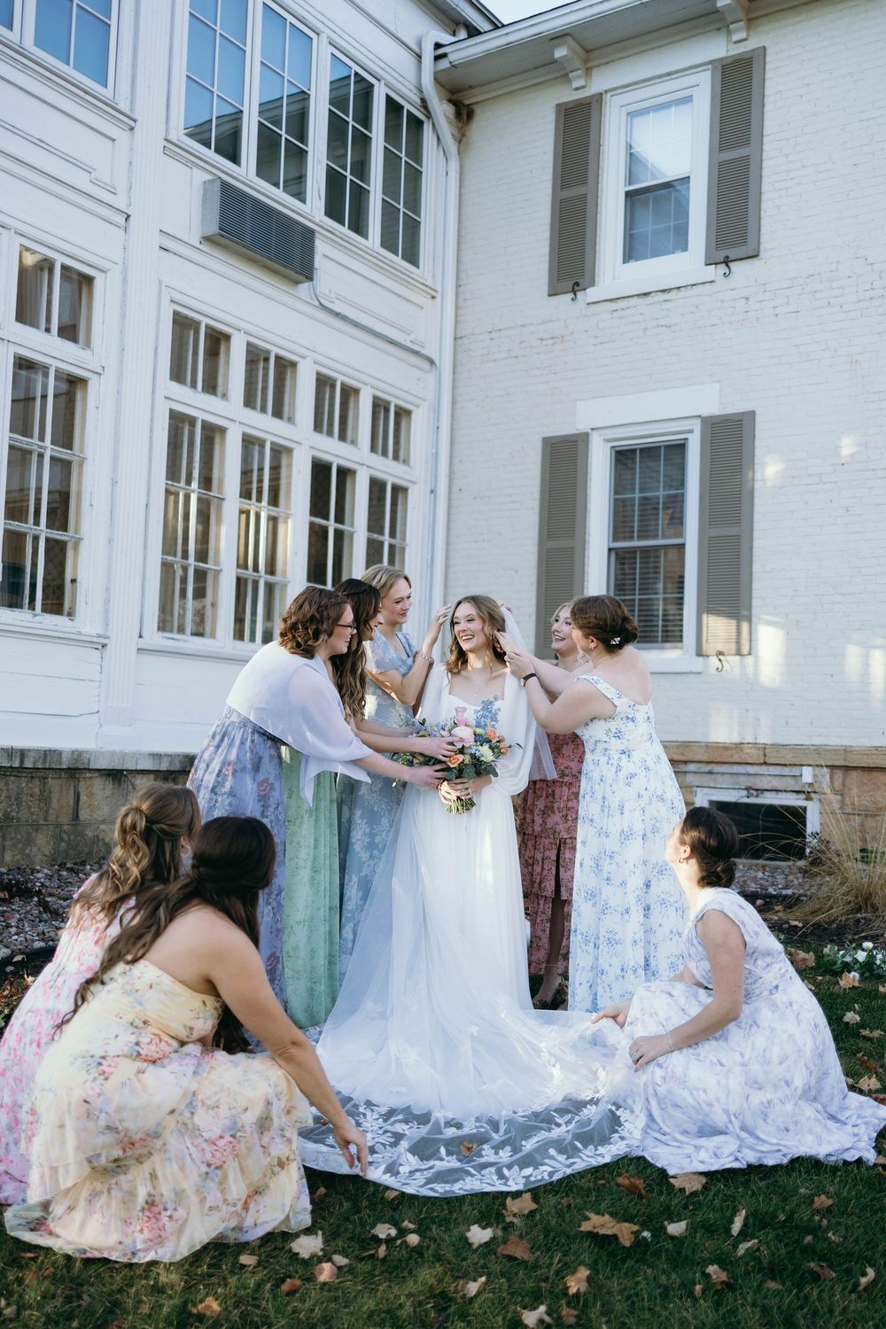 A group in pastel dresses gather on the lawn outside a white building during a wedding celebration.