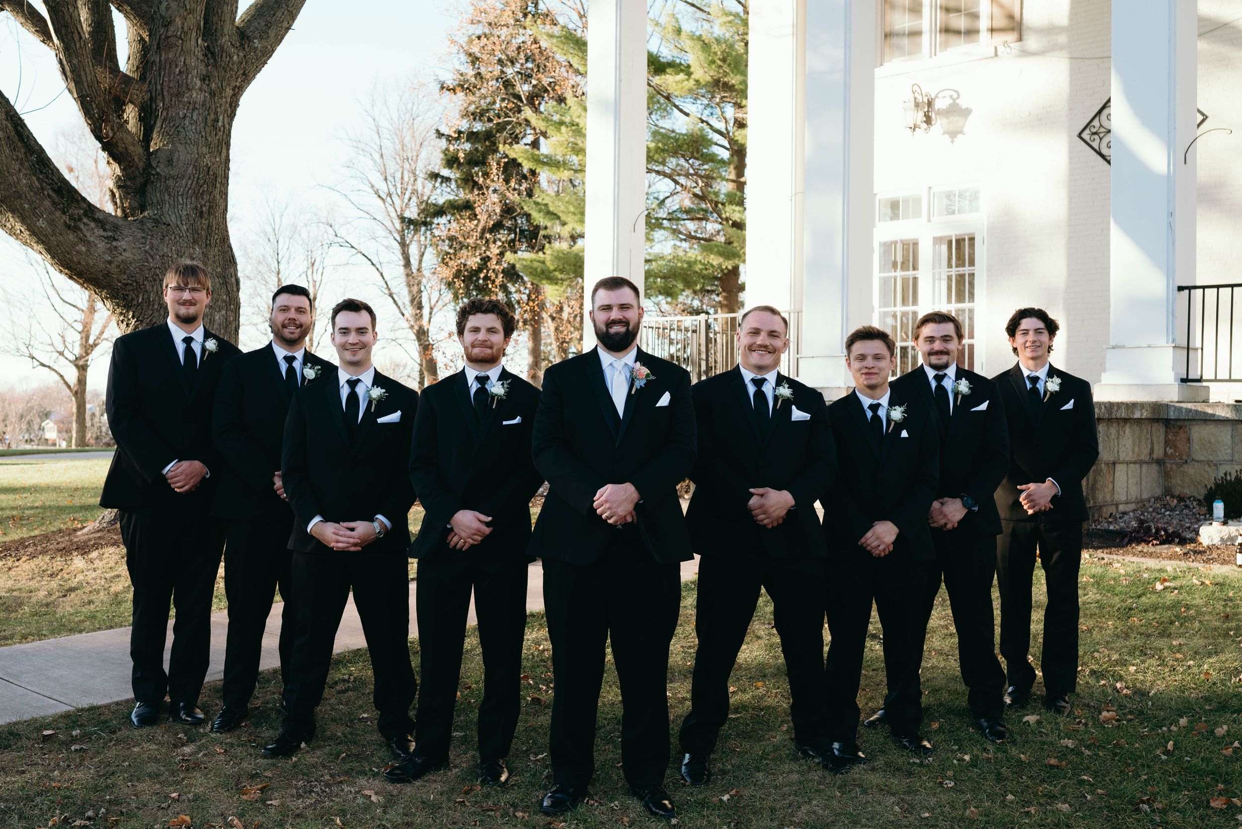 A wedding party of groomsmen in black suits and ties stand in a row outside a white building.