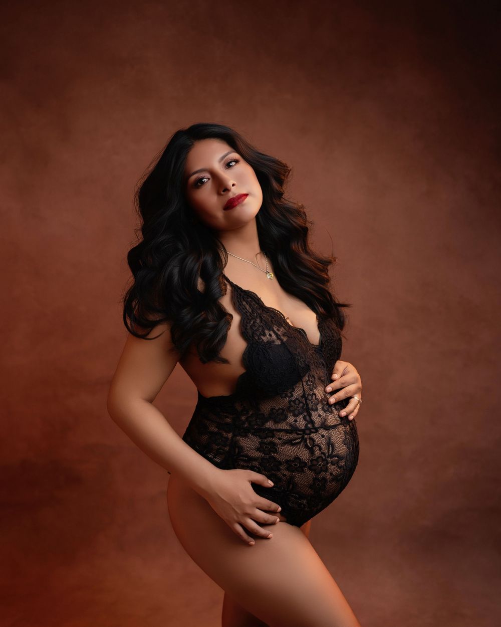 Elegant boudoir maternity photo in black lace against warm brown backdrop.
