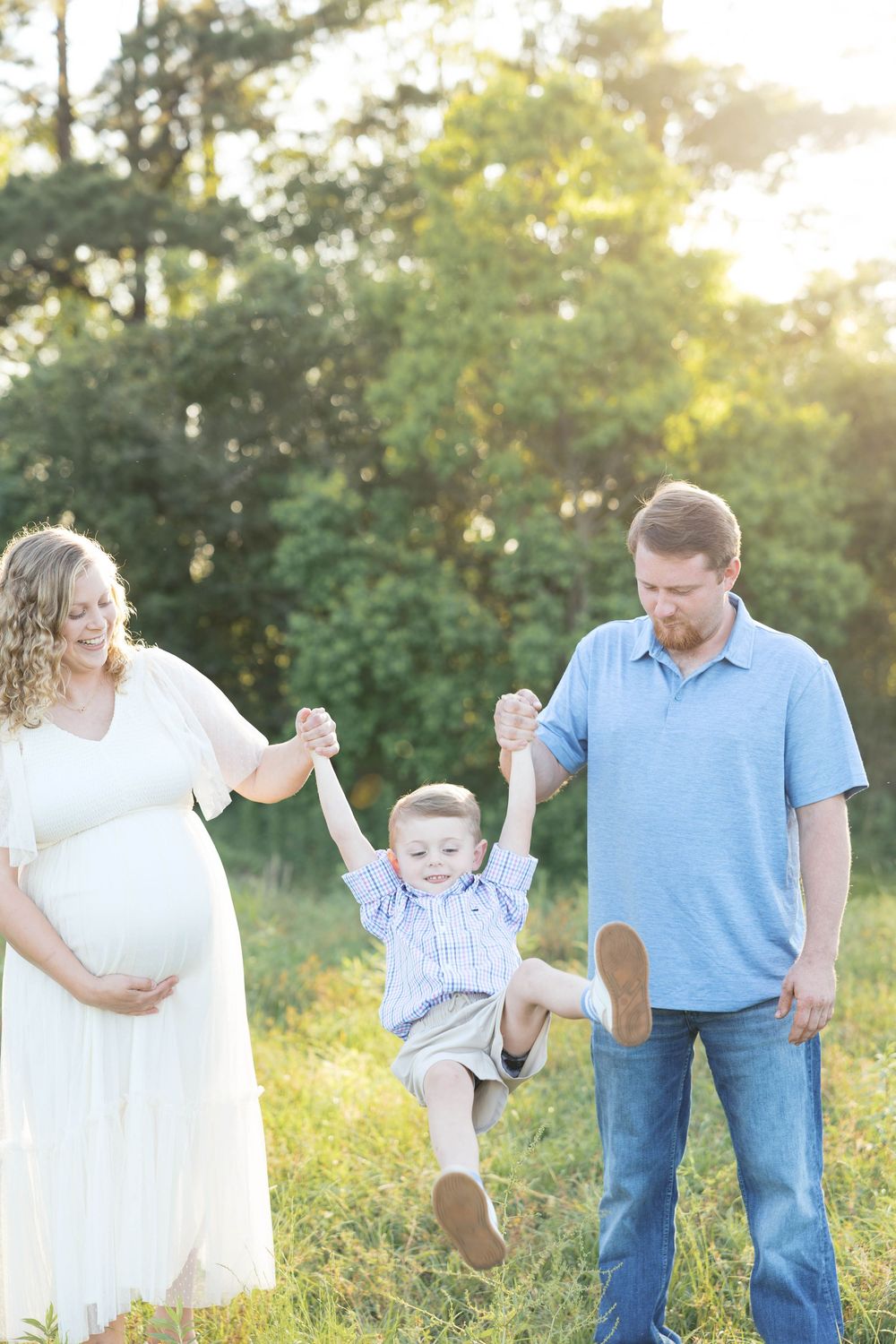 Pregnant woman in white dress and man in blue shirt swing young boy between them in sunny green field.