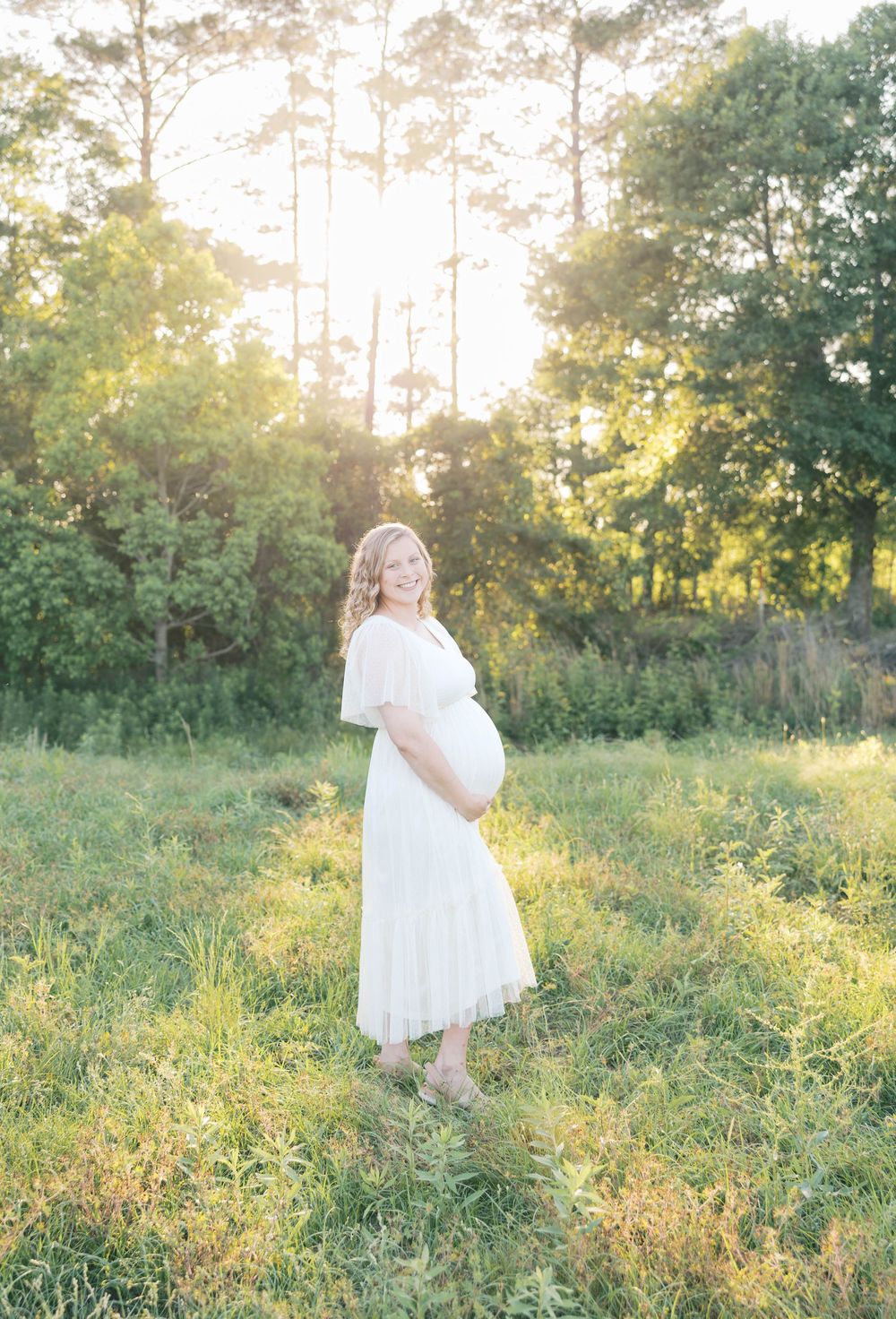 Pregnant woman in white dress standing in sunlit meadow with trees, smiling and cradling baby bump during golden hour.