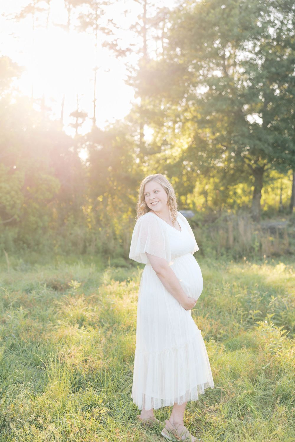Smiling pregnant woman in white dress holding baby bump outdoors in golden sunlight among green trees.