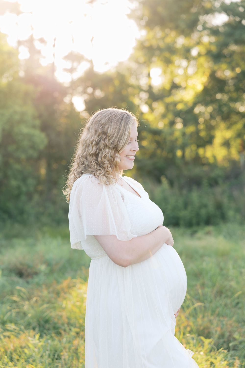 Smiling pregnant woman in white dress cradling baby bump outdoors in golden sunlight among lush green trees.