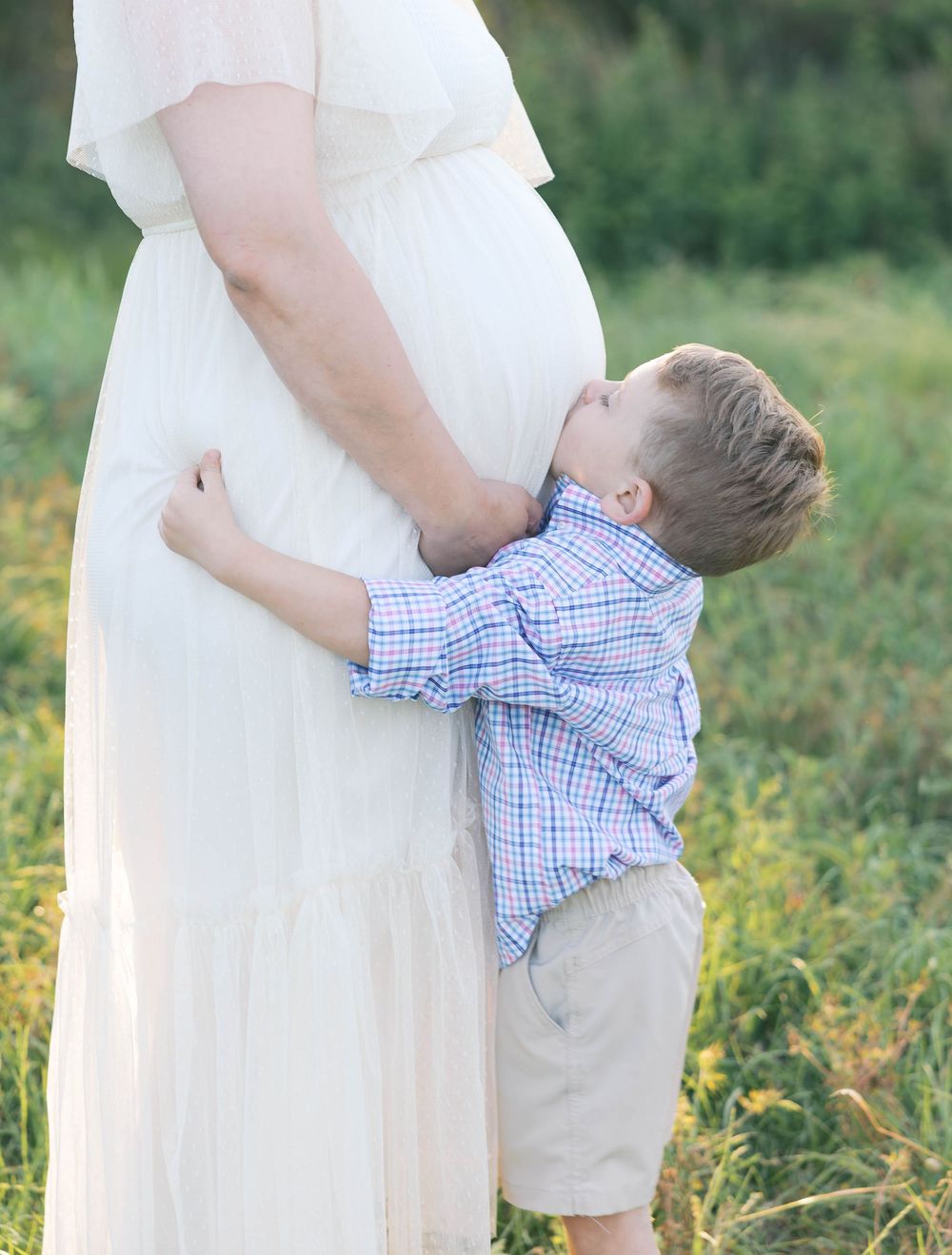 Young boy hugging pregnant mother's belly outdoors in a field during a maternity photo session.