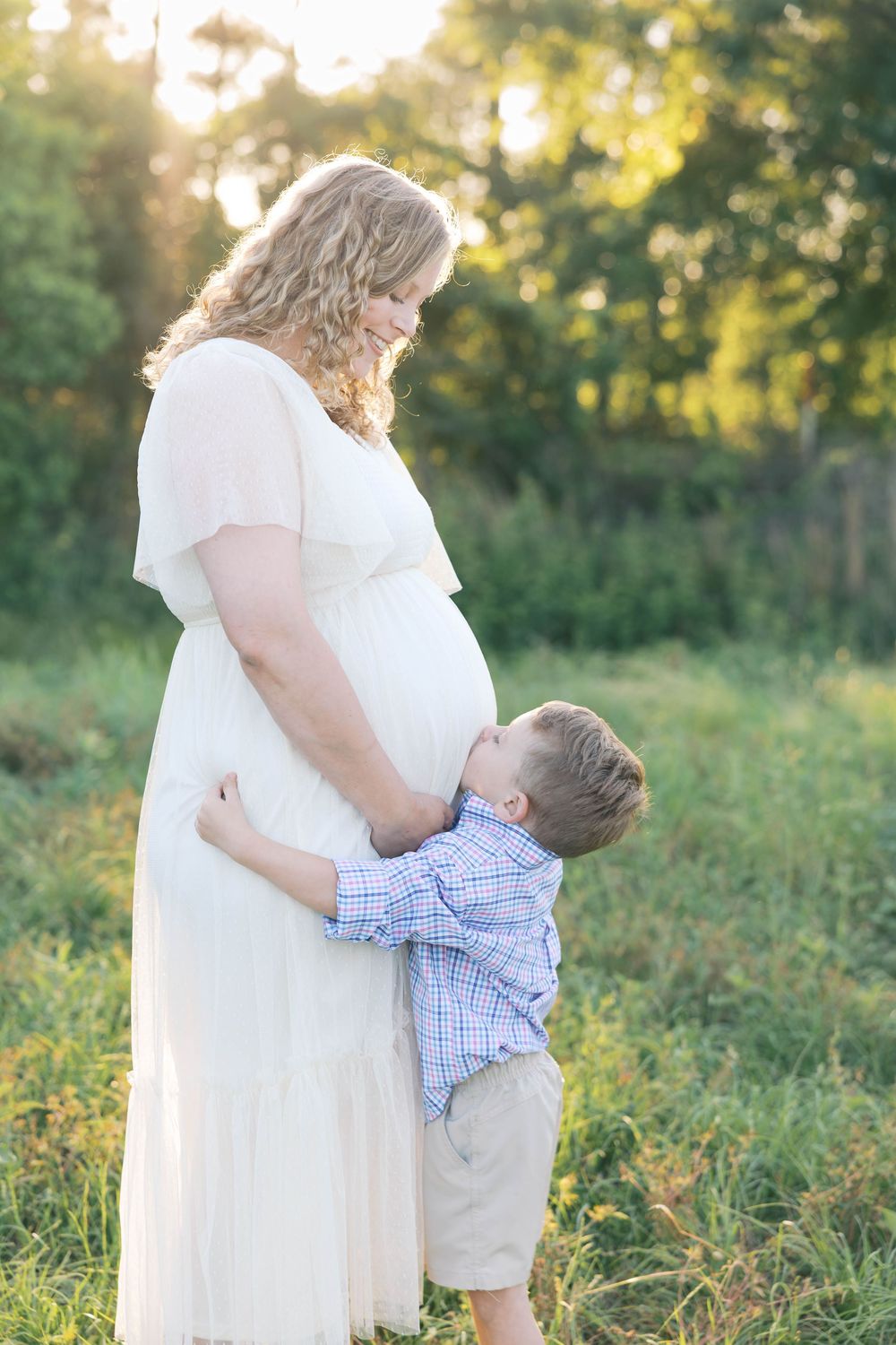 Pregnant woman in white dress outdoors with young boy hugging her baby bump in a sunlit green field.