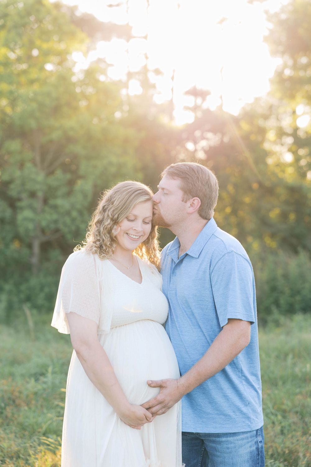 A couple shares a tender moment outdoors at golden hour, man kissing pregnant woman's temple in a sunlit green field.