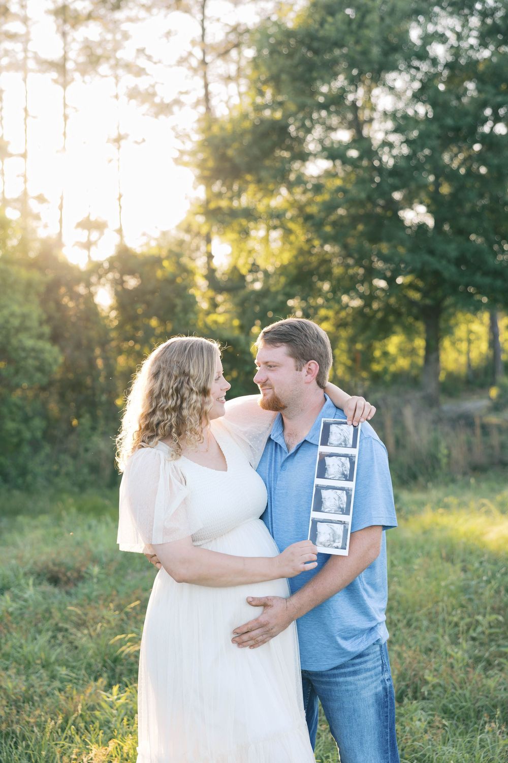 Pregnant couple smiling at each other outdoors at golden hour, man holding ultrasound photos in a sunlit field.