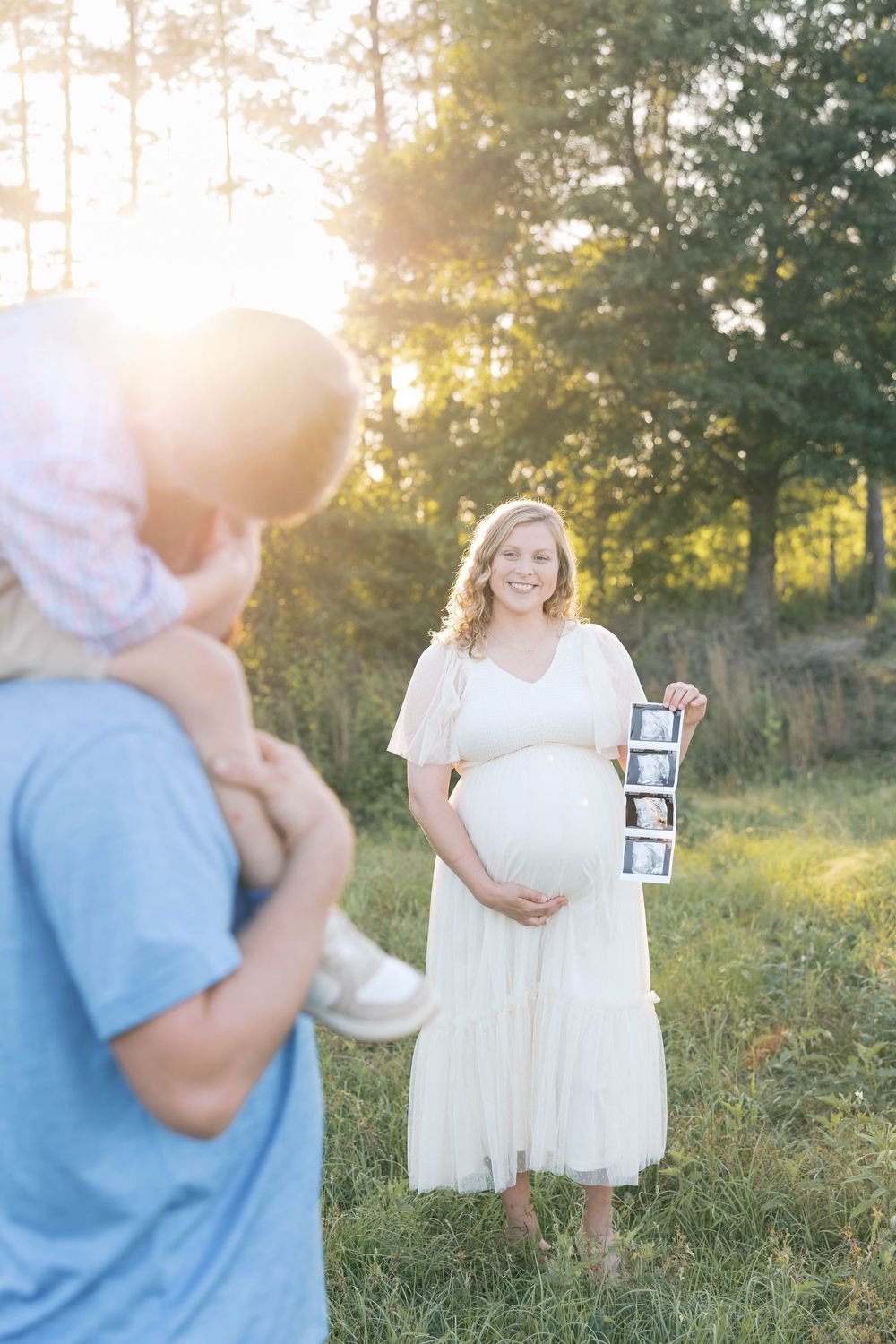 Pregnant woman in white dress holds ultrasound photos outdoors at golden hour while family watches nearby.