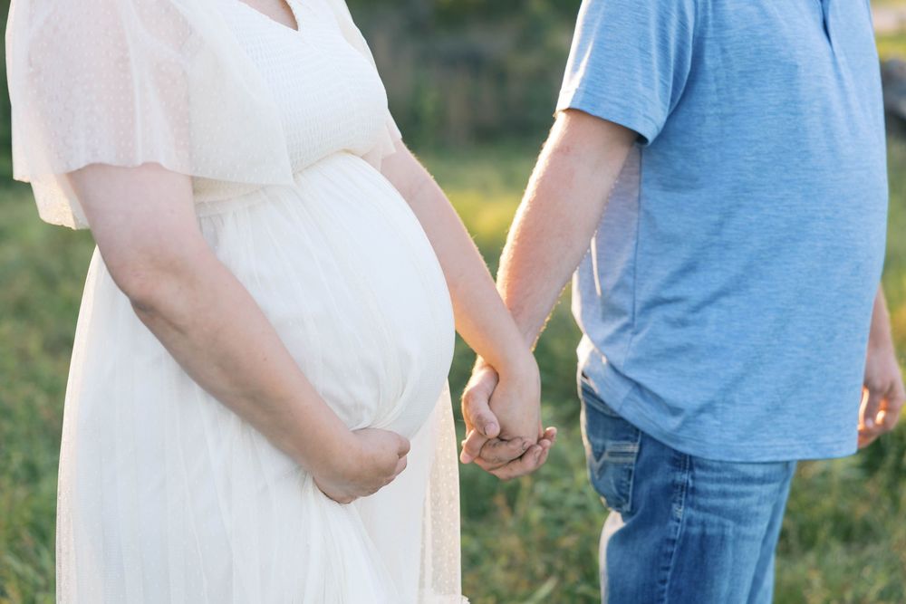 Pregnant woman in white dress holding hands with man in blue shirt outdoors in a sunny field.