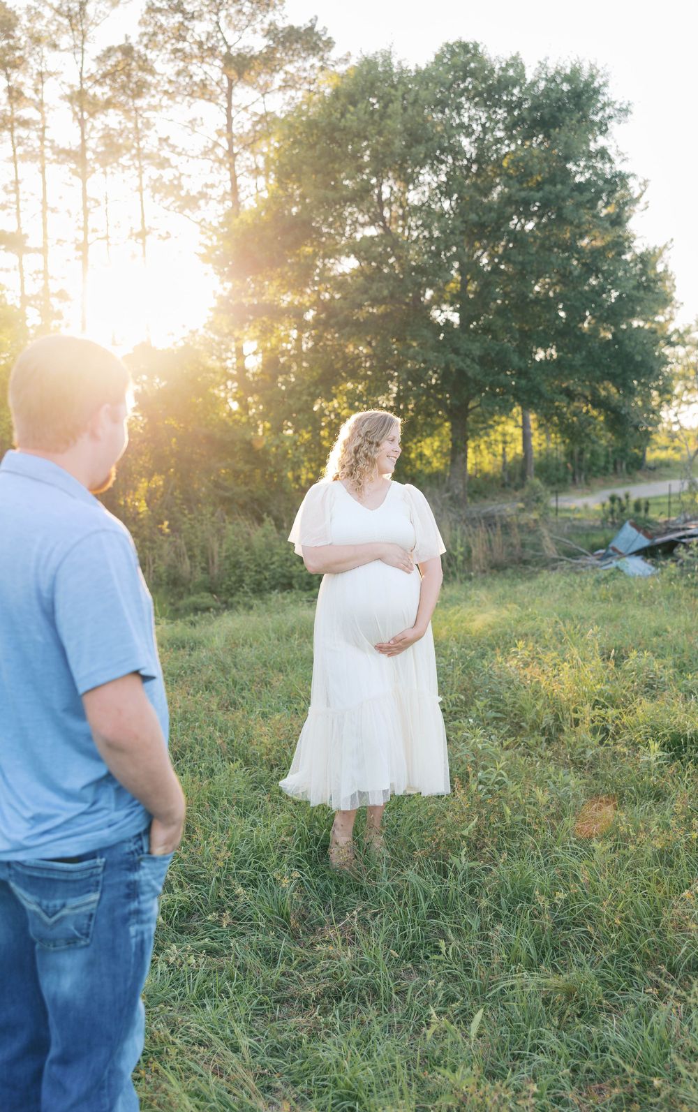 Pregnant woman in white dress holds baby bump at golden hour while man in blue shirt looks on in grassy field.