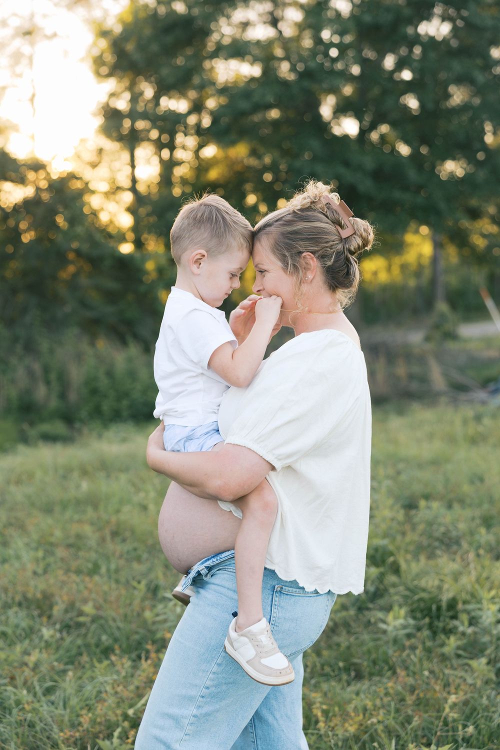 Pregnant woman in jeans holds toddler boy close, touching foreheads tenderly in a sunny outdoor setting.