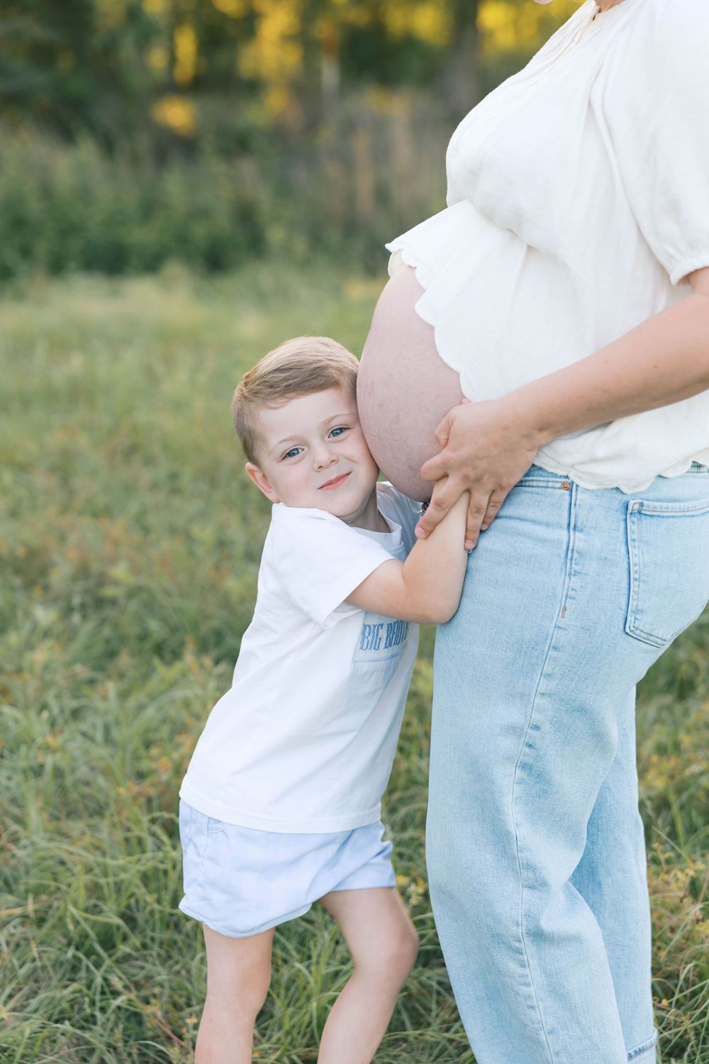 Young boy hugging pregnant mother's belly outdoors in a sunny green field, wearing white shirts in a maternity photo.