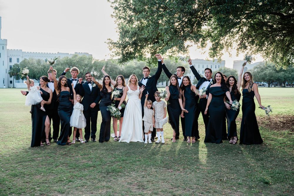 Wedding guests gather outdoors in formal attire on a grassy area for group photos.