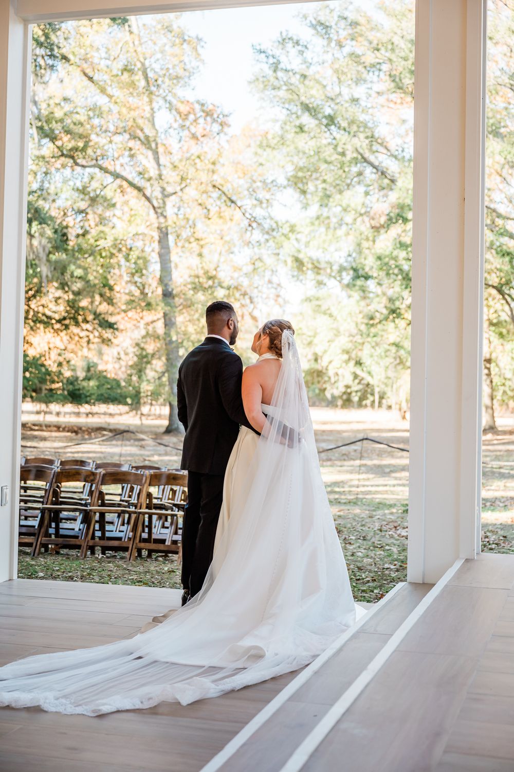 A romantic wedding moment captured in an outdoor pavilion venue with wooden chairs and fall foliage in the background.